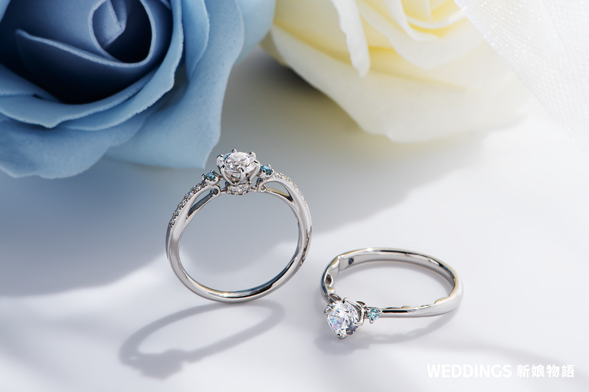 ALUXE 經典求婚鑽戒與結婚對戒放置於粉色花朵旁,展示戒台線條與鑽石光芒